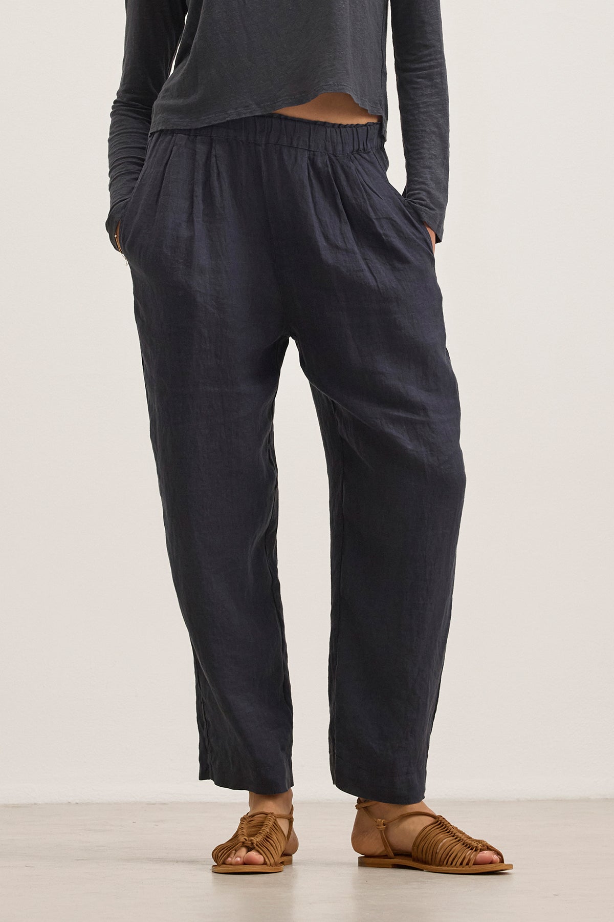 Someone wearing a pair of GINI LINEN PANTS by Velvet by Graham & Spencer stands on a light-colored floor. The dark, loose-fitting pants with an elastic waistband, featuring a straight-leg silhouette, pair effortlessly with open-toed sandals for a relaxed yet stylish look.