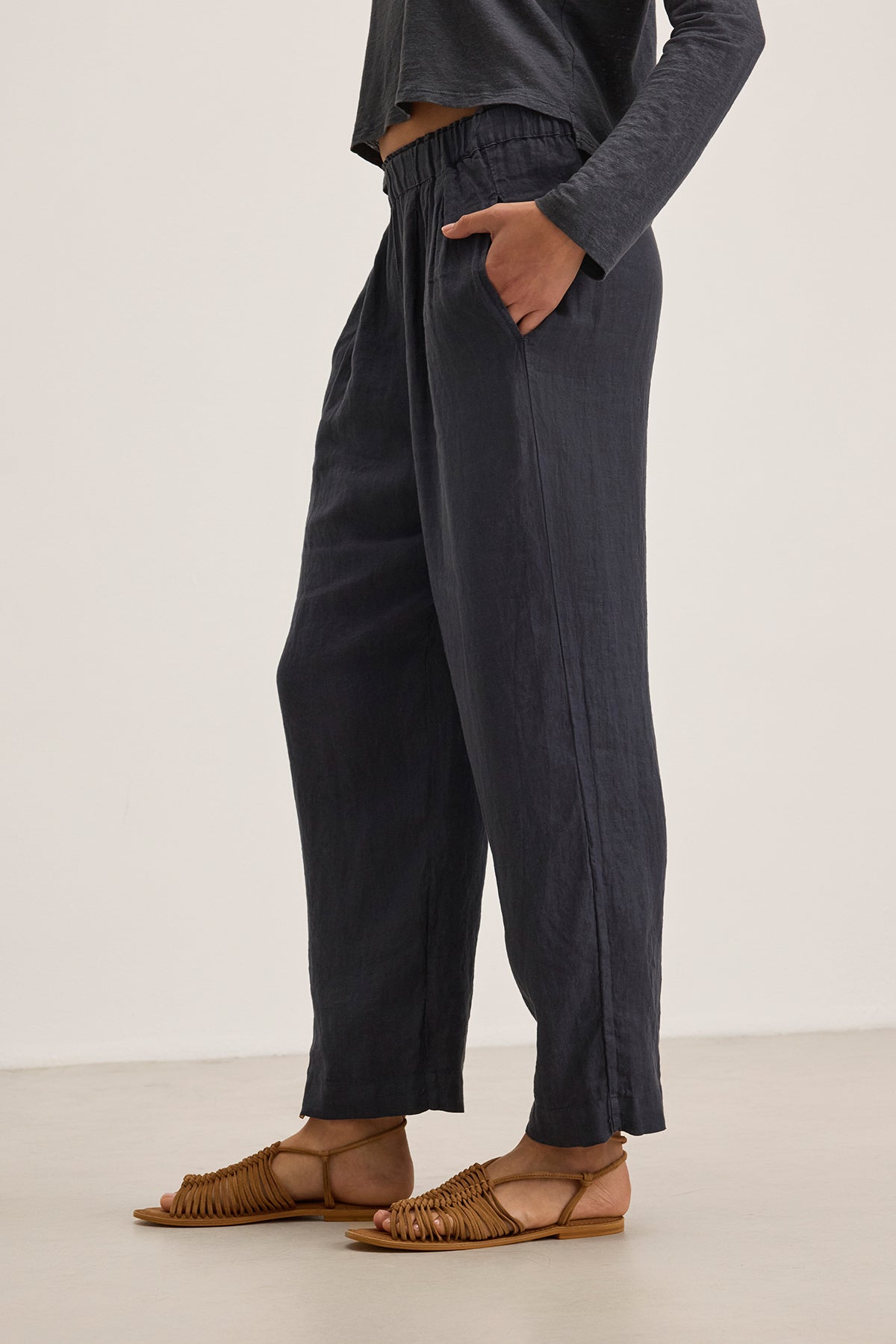 A person stands on a light surface wearing Velvet by Graham & Spencer's GINI LINEN PANT, which features a straight-leg silhouette for a relaxed look, paired with an elastic-waistband dark long-sleeve shirt and brown woven sandals.