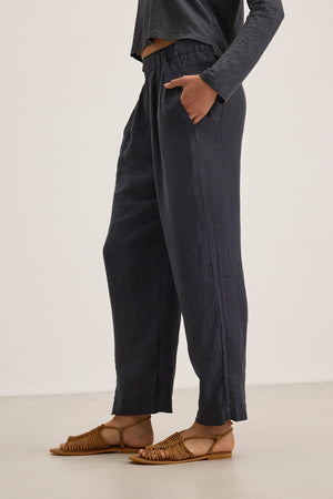 A person stands on a light surface wearing Velvet by Graham & Spencer's GINI LINEN PANT, which features a straight-leg silhouette for a relaxed look, paired with an elastic-waistband dark long-sleeve shirt and brown woven sandals.