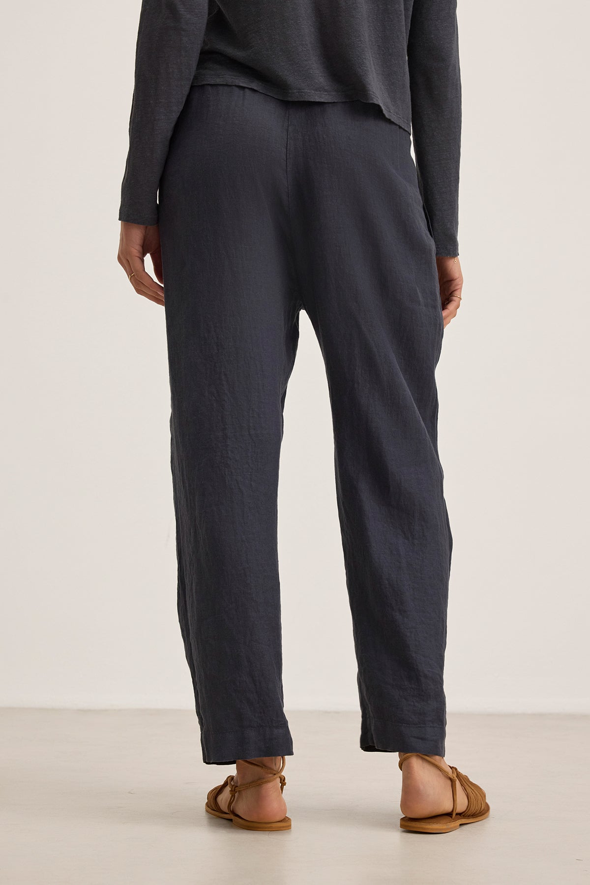 Facing away from the camera, a person wears dark gray GINI LINEN PANTs and a matching top from Velvet by Graham & Spencer. Brown sandals complete this effortlessly chic look.