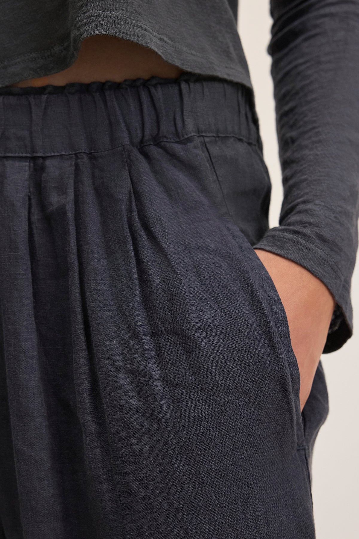 A person wearing a dark long-sleeve shirt and Velvet by Graham & Spencer's GINI LINEN PANT, featuring a straight-leg silhouette and pleats, stands with hands in pockets.