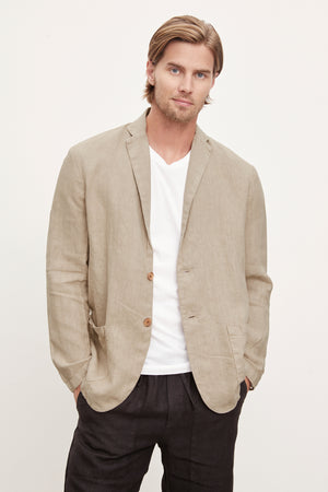 A man with long blond hair wearing a Velvet by Graham & Spencer JOSHUA LINEN BLAZER over a white t-shirt, paired with dark trousers, standing against a light background.