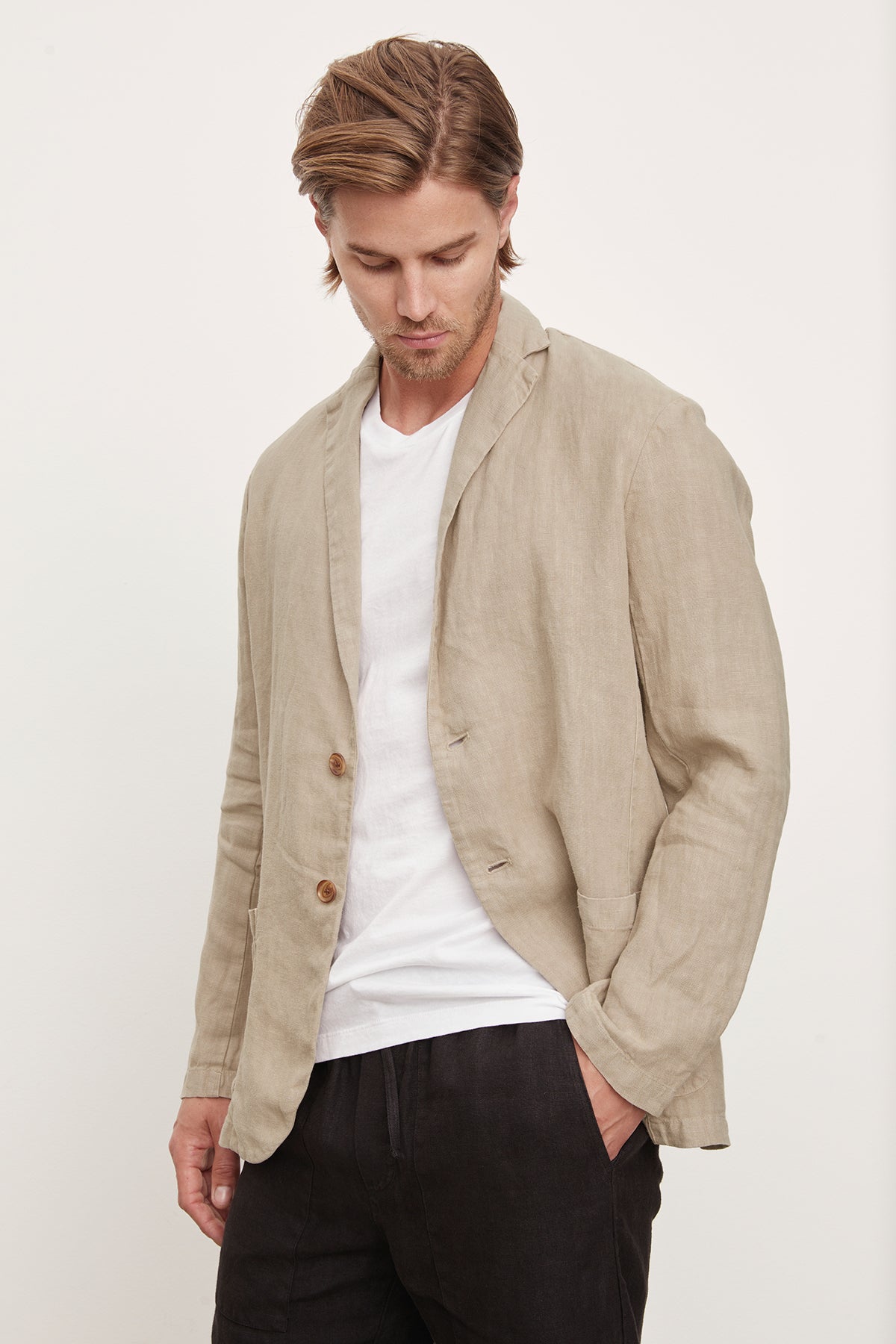 Man wearing a beige Joshua Linen Blazer by Velvet by Graham & Spencer over a white t-shirt, paired with Phelan Linen Pants, looking away thoughtfully.