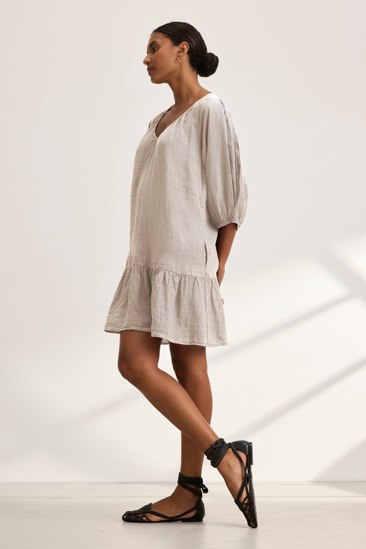 A woman wearing the KAYLIE LINEN DRESS by Velvet by Graham & Spencer stands against a plain white background. The dress features a light, woven linen fabric and a tiered silhouette. She completes her look with black sandals and styled hair in a bun.-38943210602689