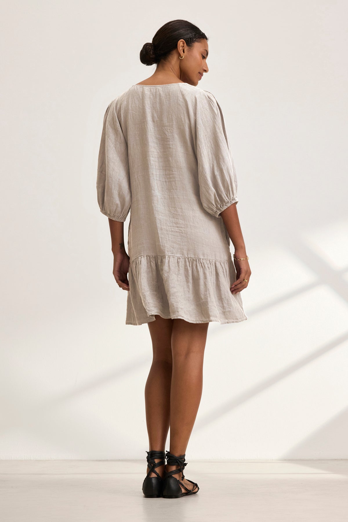 A barefoot woman stands with her back to the camera in a well-lit space, wearing the KAYLIE LINEN DRESS by Velvet by Graham & Spencer, a light beige woven linen dress with balloon sleeves and a graceful, tiered silhouette.-38943210733761