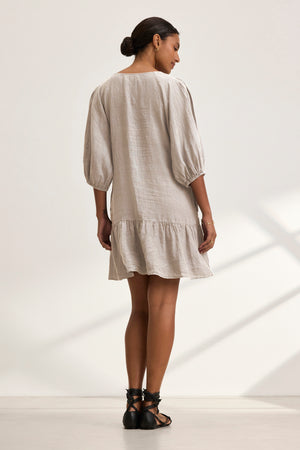 A barefoot woman stands with her back to the camera in a well-lit space, wearing the KAYLIE LINEN DRESS by Velvet by Graham & Spencer, a light beige woven linen dress with balloon sleeves and a graceful, tiered silhouette.
