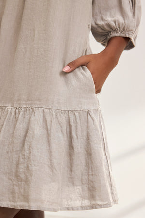 Close-up of a person wearing Velvet by Graham & Spencer's KAYLIE LINEN DRESS, a beige loose-fitting woven linen garment with puff sleeves and a ruffled hem. The tiered silhouette adds elegance as manicured nails in light pink rest inside the side pocket.