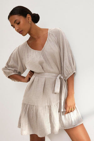 A woman models the KAYLIE LINEN DRESS by Velvet by Graham & Spencer, featuring light beige fabric, puffed long sleeves, and a belt, against a white backdrop.