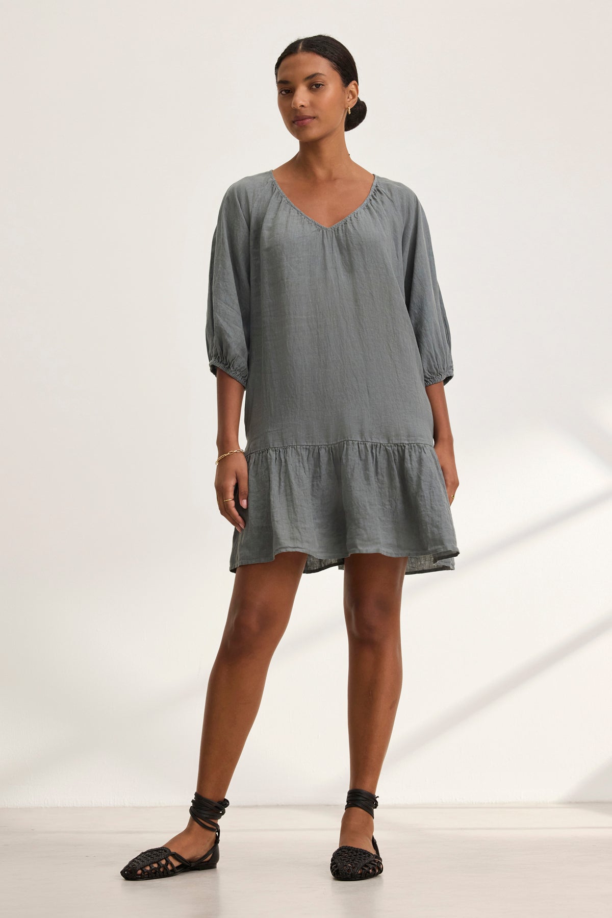 A woman wears the KAYLIE LINEN DRESS by Velvet by Graham & Spencer, featuring a gray woven linen style with elbow-length puff sleeves and a ruffled hem, paired with black strappy flats.-38943210537153