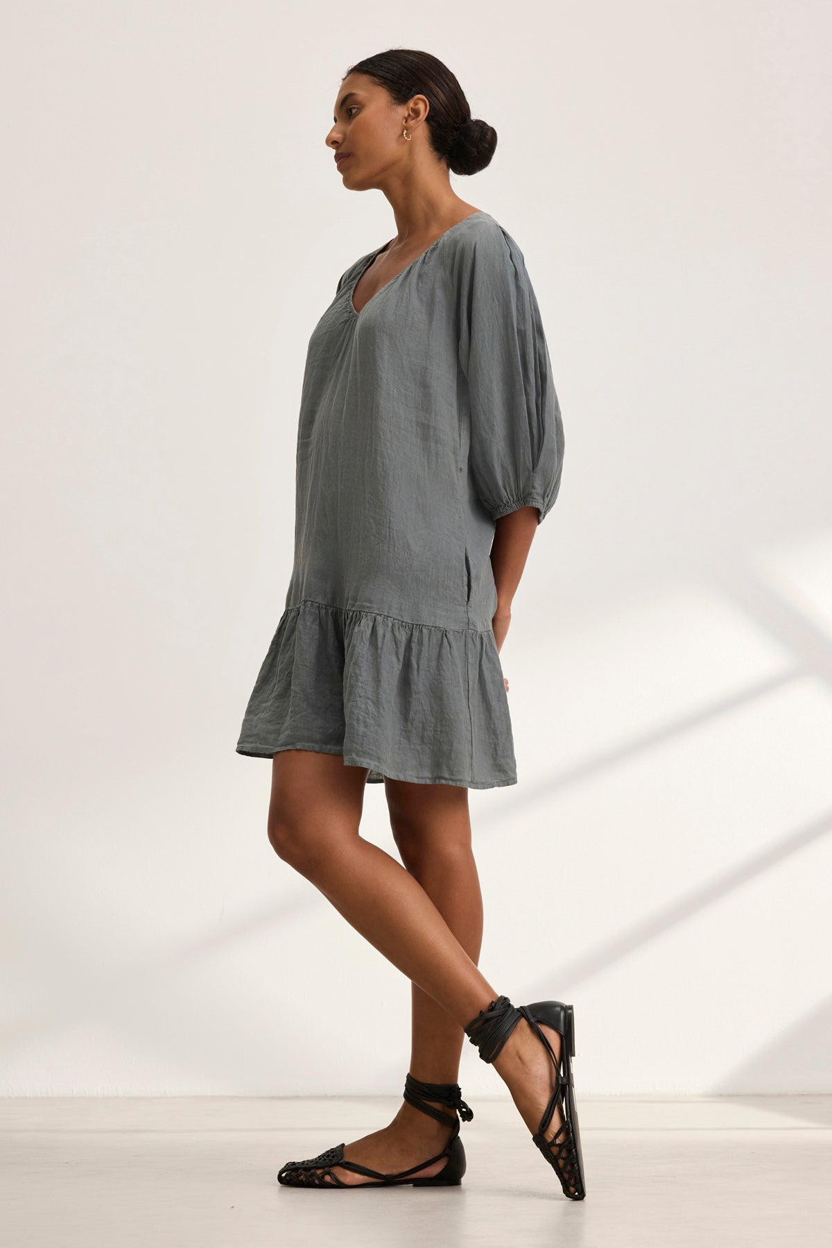 A woman in the KAYLIE LINEN DRESS by Velvet by Graham & Spencer stands sideways, wearing black strappy shoes. Her hair is tied back, and she's in a bright, minimalist room.-38943210504385