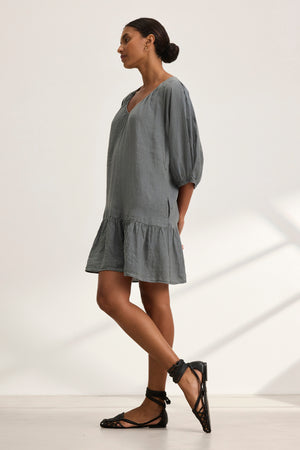 A woman in the KAYLIE LINEN DRESS by Velvet by Graham & Spencer stands sideways, wearing black strappy shoes. Her hair is tied back, and she's in a bright, minimalist room.