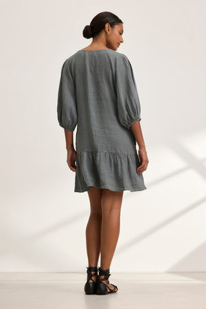 A person stands with their back turned, wearing the KAYLIE LINEN DRESS by Velvet by Graham & Spencer. The gray woven linen dress features puffed sleeves and a tiered silhouette. Their hair is tied up, as gentle light from the window casts intricate shadows on the floor.