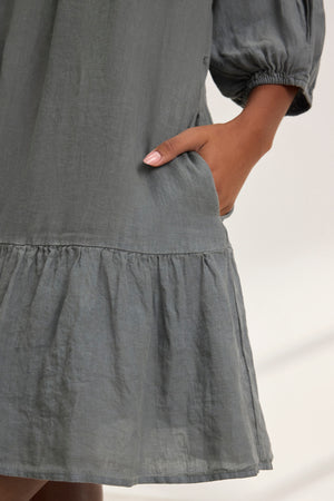 A person wears the KAYLIE LINEN DRESS by Velvet by Graham & Spencer, featuring a gray woven linen fabric with puff sleeves and a tiered silhouette. Their hand rests comfortably in the pocket.