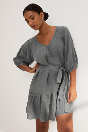 A woman models the Velvet by Graham & Spencer KAYLIE LINEN DRESS, featuring a gray woven linen fabric, tiered silhouette, tied waist, and three-quarter puff sleeves, against a plain white background.