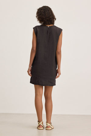 A person with curly hair stands facing away in a sleeveless LARA LINEN DRESS by Velvet by Graham & Spencer and white sandals, against a plain white background.