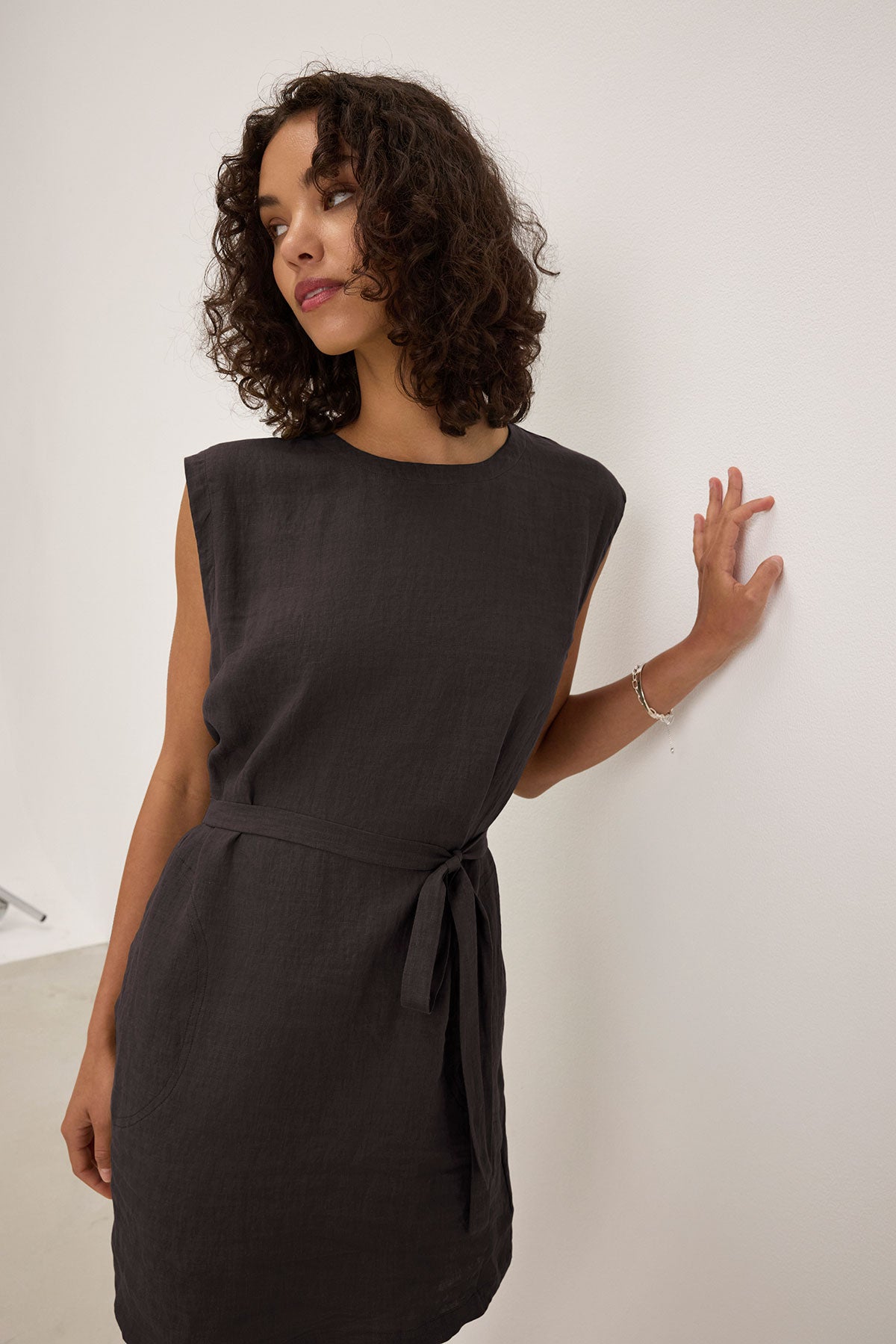 A person with curly hair wearing a sleeveless LARA LINEN DRESS by Velvet by Graham & Spencer stands against a white wall, looking to the side.