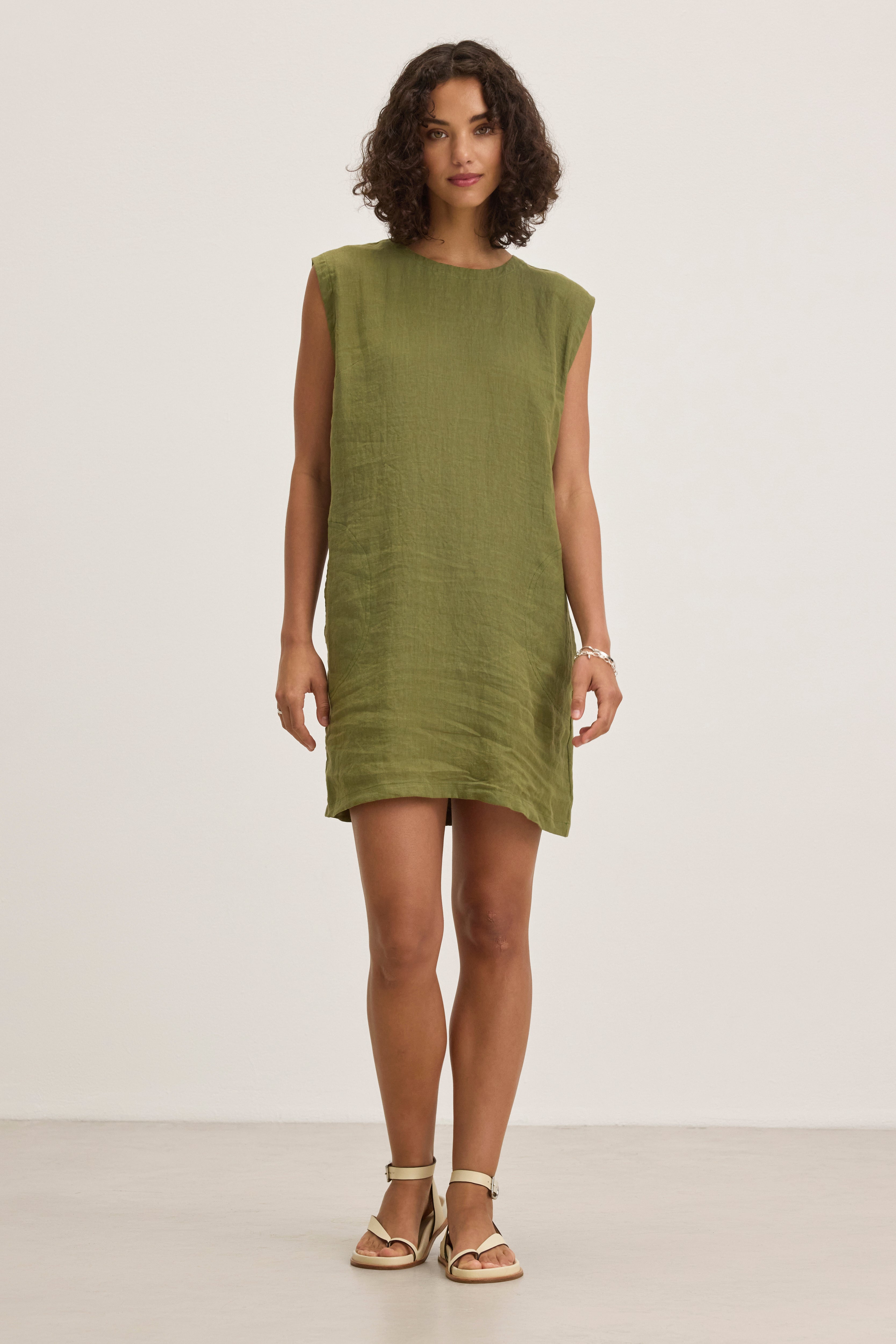 A woman stands in a plain room wearing the LARA LINEN DRESS by Velvet by Graham & Spencer, featuring a sleeveless design and detachable belt, paired with light-colored sandals and looking straight ahead with a neutral expression.