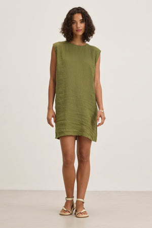 A woman stands in a plain room wearing the LARA LINEN DRESS by Velvet by Graham & Spencer, featuring a sleeveless design and detachable belt, paired with light-colored sandals and looking straight ahead with a neutral expression.