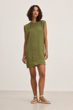 Someone wears the LARA LINEN DRESS by Velvet by Graham & Spencer, a sleeveless green piece, standing against a plain background. They have short curly hair and wear a bracelet and sandals.