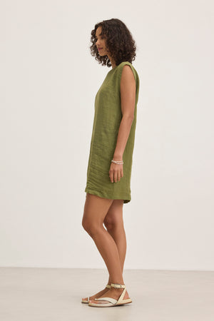 A person with curly hair is standing sideways, wearing the LARA LINEN DRESS by Velvet by Graham & Spencer and sandals against a plain background.