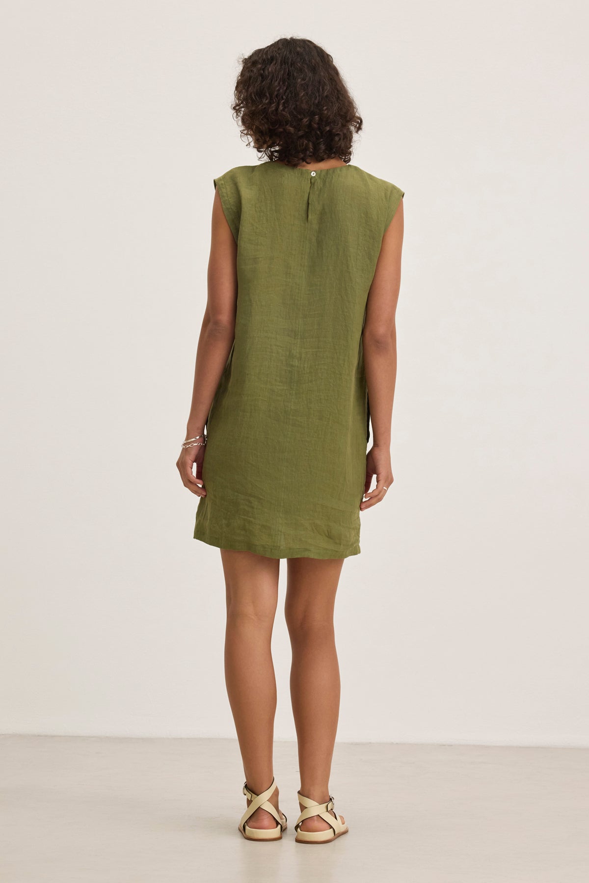 A person stands against a plain background, wearing the LARA LINEN DRESS by Velvet by Graham & Spencer—a sleeveless green dress—paired with sandals. They have curly hair and face away from the camera.