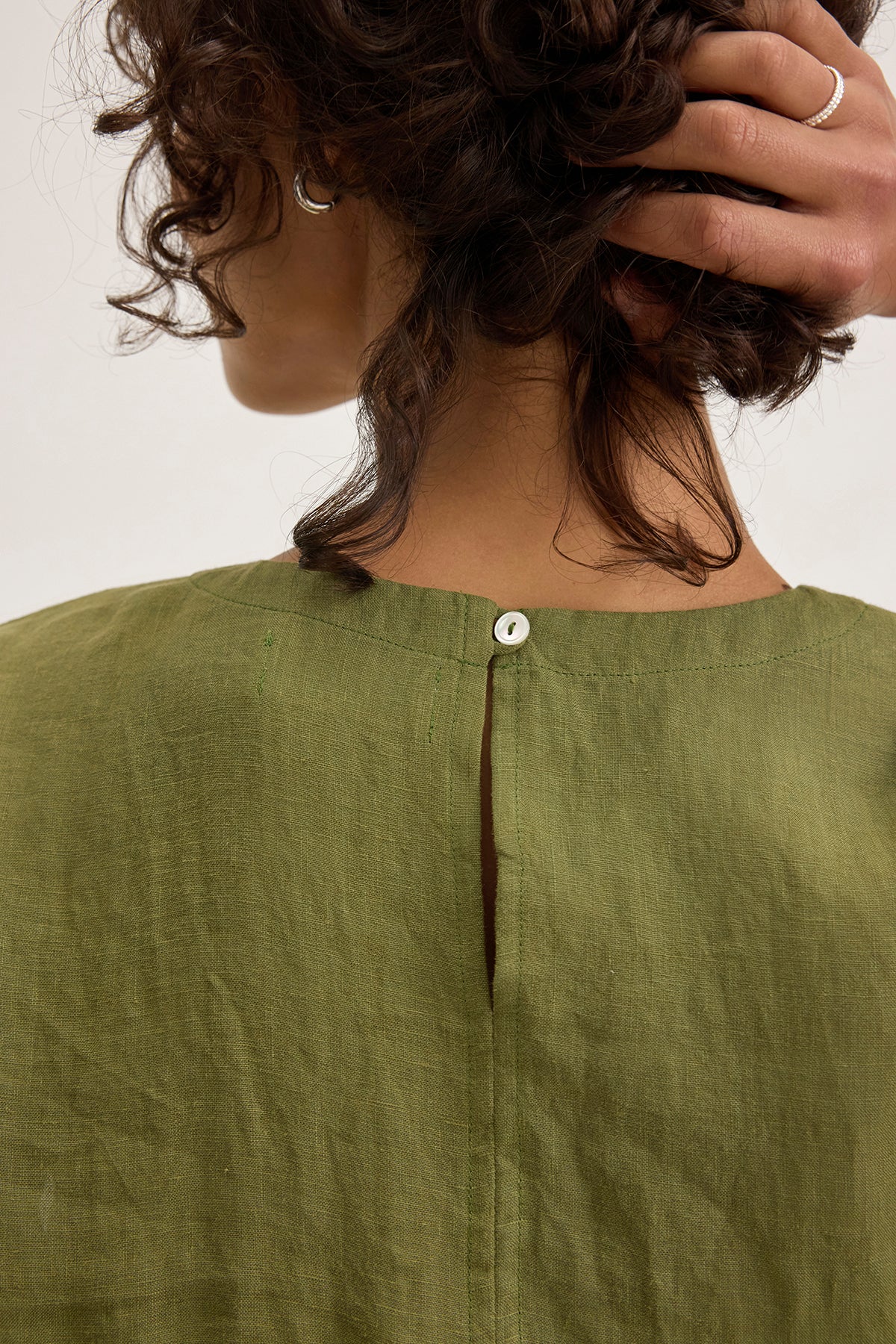 A person with curly brown hair holds their head, showing the back of the Velvet by Graham & Spencer LARA LINEN DRESS in green, which features a small white button at the neckline and a detachable belt.