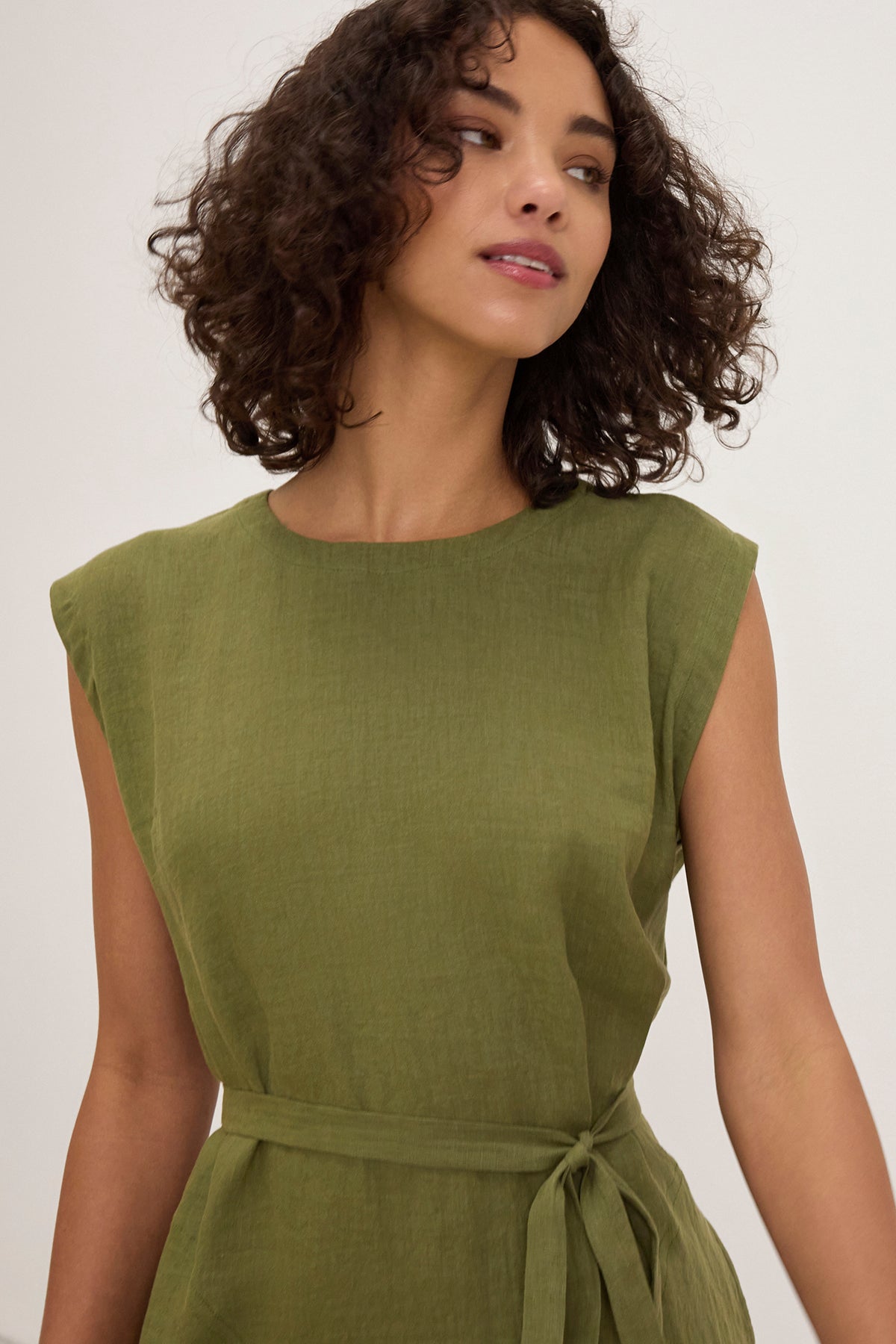 A woman with curly hair wears the LARA LINEN DRESS by Velvet by Graham & Spencer, an olive green, sleeveless linen dress with a detachable belt, standing against a plain background.
