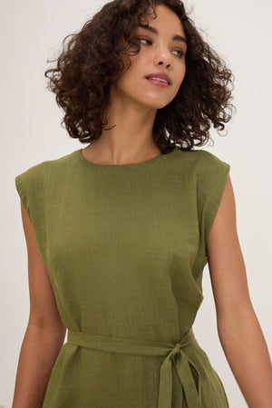 A woman with curly hair wears the LARA LINEN DRESS by Velvet by Graham & Spencer, an olive green, sleeveless linen dress with a detachable belt, standing against a plain background.