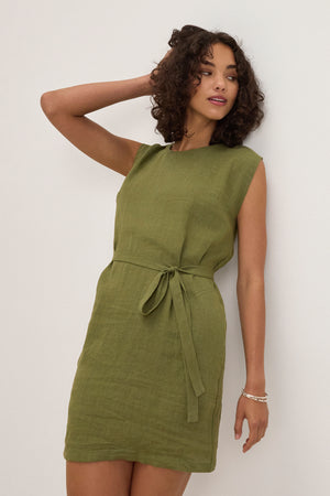 A woman poses against a white wall in the LARA LINEN DRESS by Velvet by Graham & Spencer, an olive sleeveless linen dress with a detachable belt, her arm bent and hand touching her head.