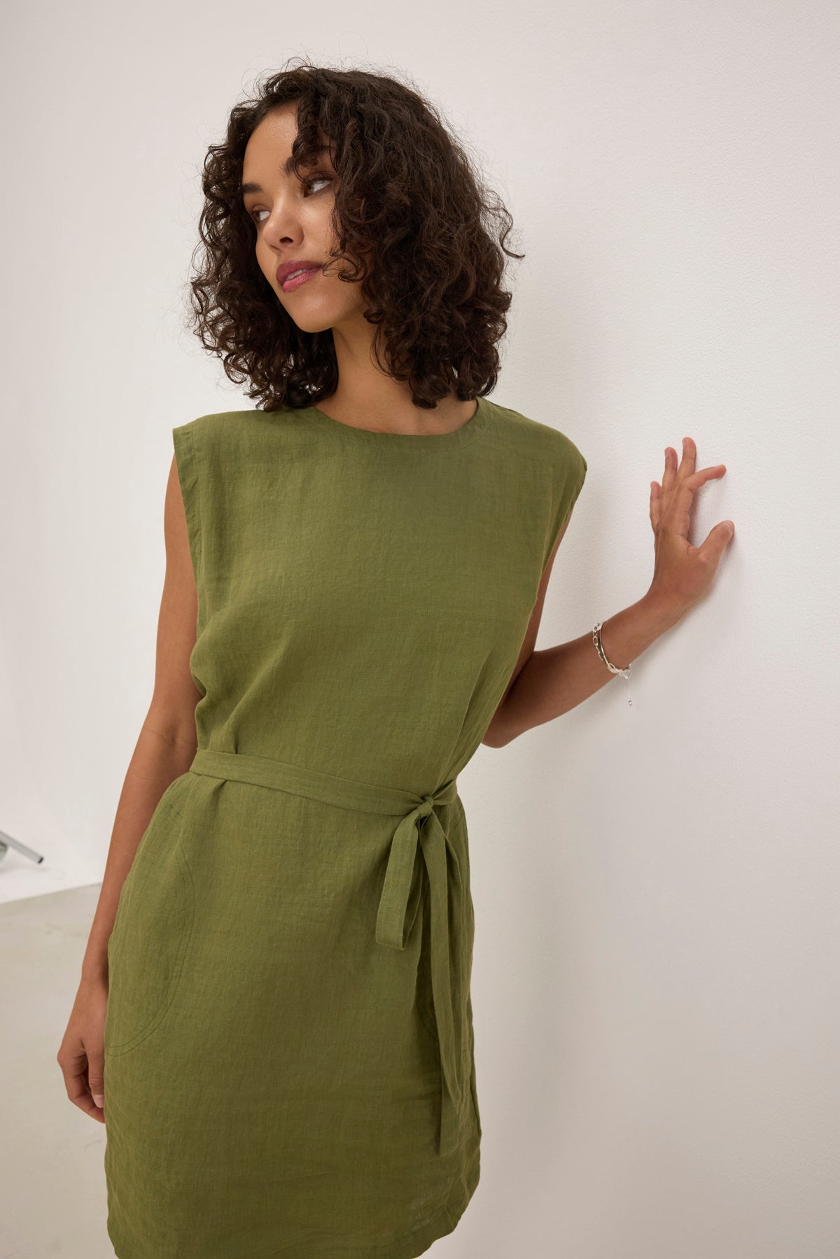 A person wearing a LARA LINEN DRESS by Velvet by Graham & Spencer, featuring a tied waist belt, stands against a white wall while looking to the side.