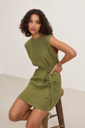 A woman with curly hair wears the Velvet by Graham & Spencer LARA LINEN DRESS, featuring a sleeveless design and detachable belt, while seated on a wooden stool in a minimalist room.
