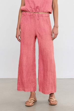 A person wearing lightweight pink LOLA LINEN PANT by Velvet by Graham & Spencer with an elastic waistband and drawstring, a matching top, a bracelet on the left wrist, and beige sandals on a plain grey floor.