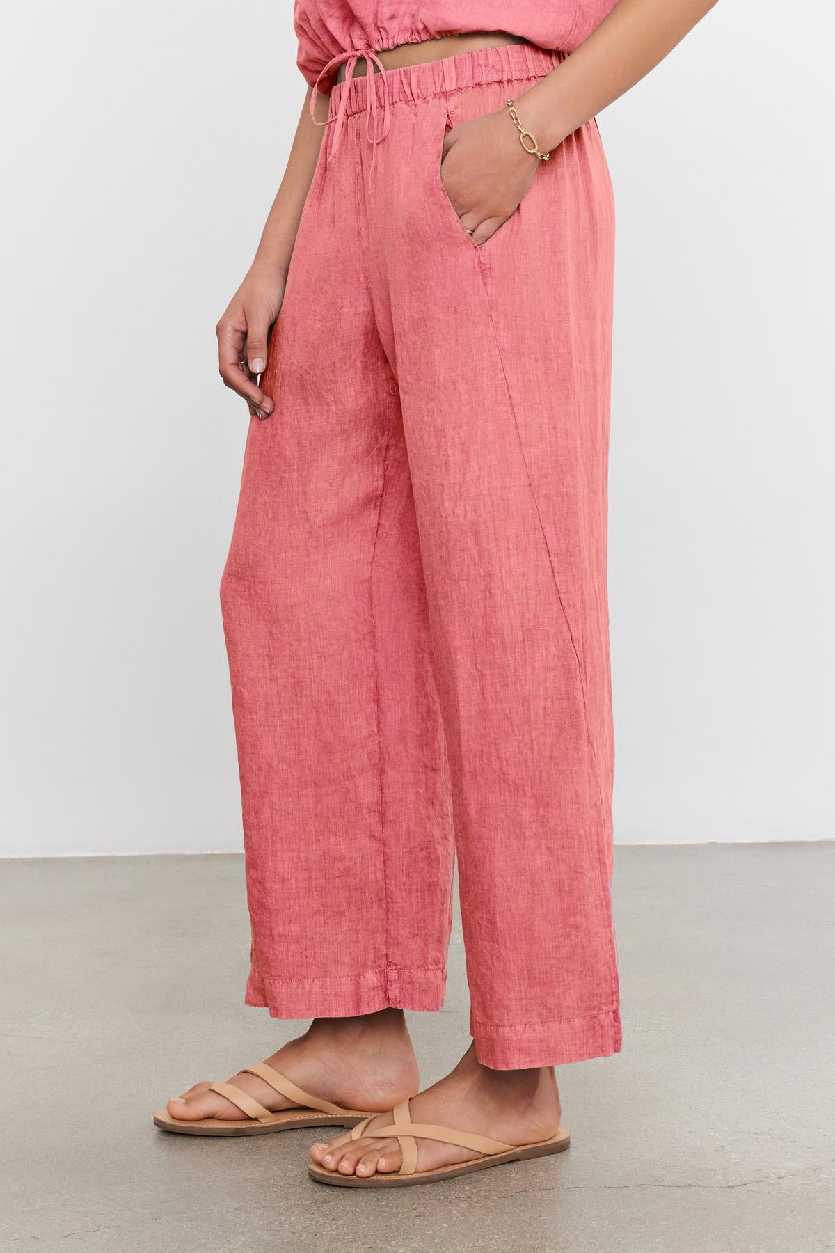 A person is wearing loose-fitting pink LOLA LINEN PANT by Velvet by Graham & Spencer with hands in pockets, a matching top, and beige sandals.