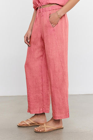 A person is wearing loose-fitting pink LOLA LINEN PANT by Velvet by Graham & Spencer with hands in pockets, a matching top, and beige sandals.