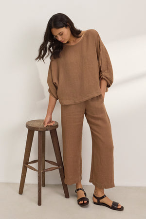 A woman stands by a wooden stool against a white wall, wearing the Velvet by Graham & Spencer LOLA LINEN PANT—a lightweight, brown, pull-on trouser with an elastic waist—paired with a loose long-sleeve top.