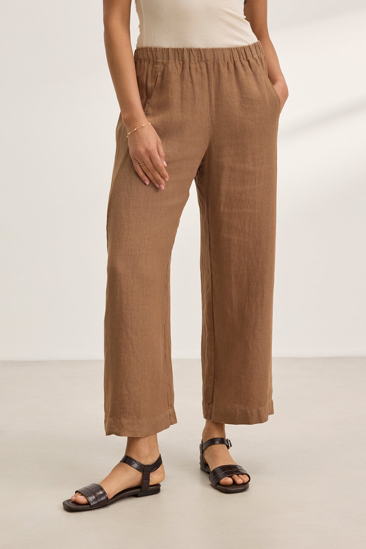 Someone wearing Velvet by Graham & Spencer's LOLA LINEN PANT with a relaxed leg, a white tank top, and black sandals, standing on a light-colored floor.-38662940852417