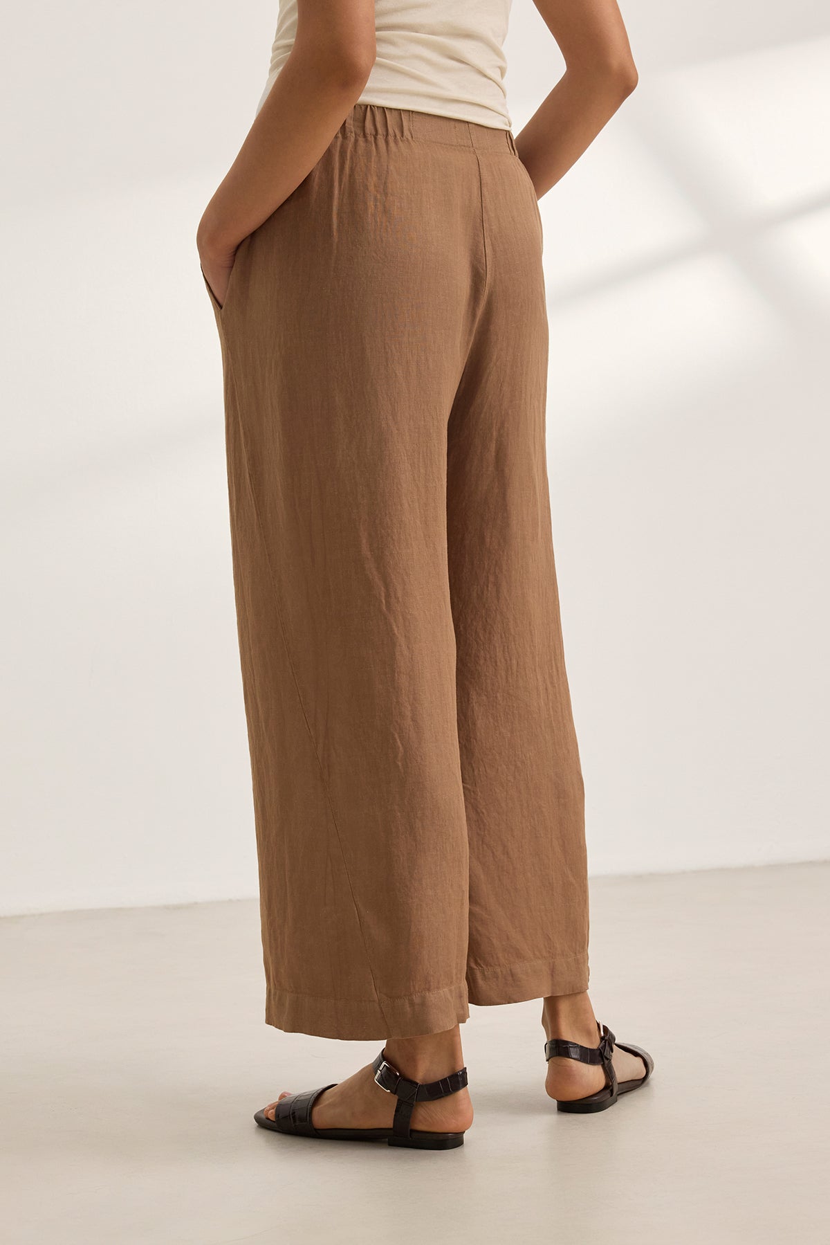 A person wearing the LOLA LINEN PANT by Velvet by Graham & Spencer—brown with an elastic waist—and black sandals stands on a light-colored floor, hands in pockets.