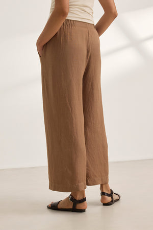 A person wearing the LOLA LINEN PANT by Velvet by Graham & Spencer—brown with an elastic waist—and black sandals stands on a light-colored floor, hands in pockets.