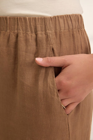 Close-up of a person wearing Velvet by Graham & Spencer's LOLA LINEN PANT in brown, featuring an elastic waist and casually resting one hand in the side pocket.