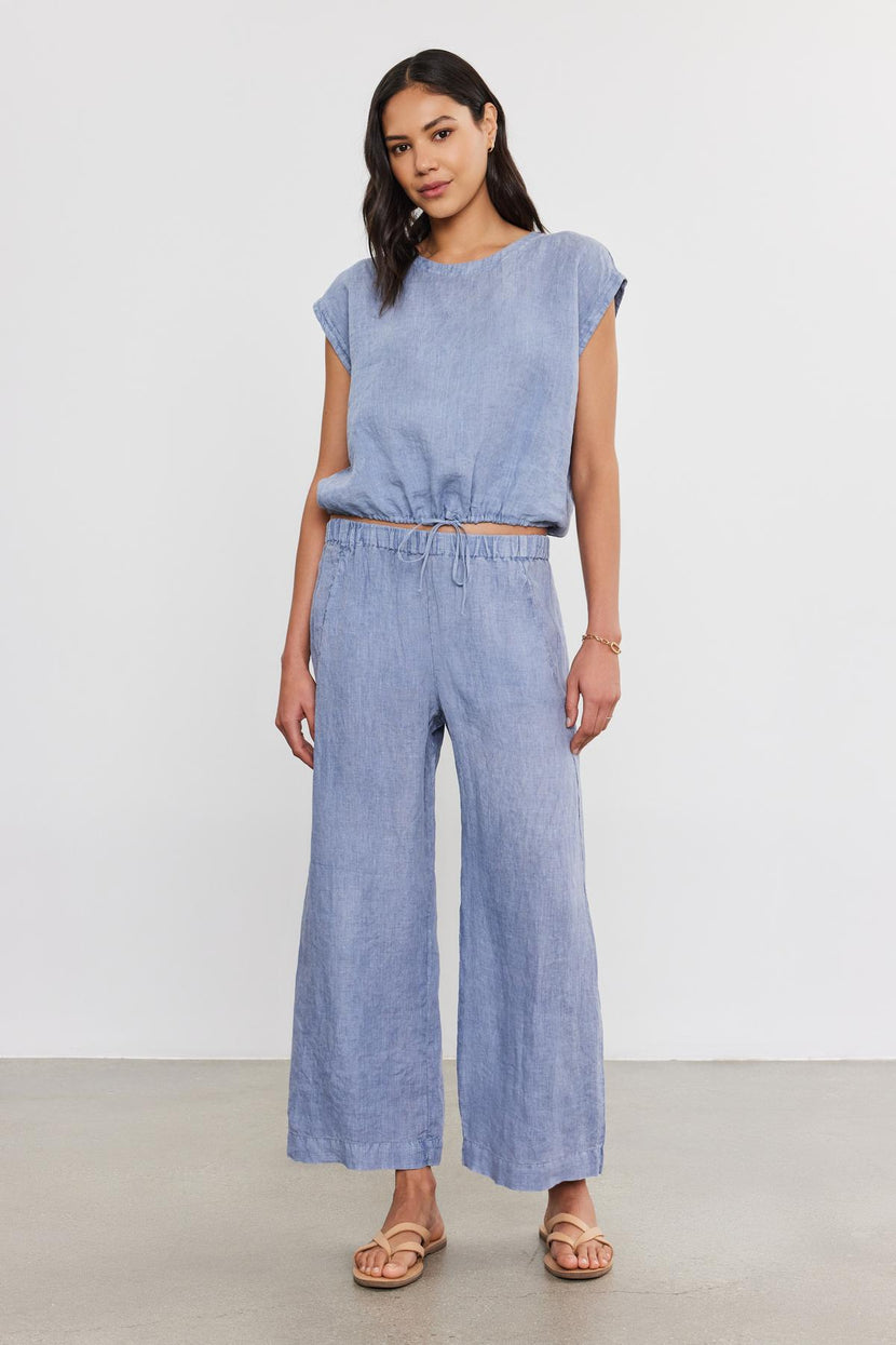 A woman in a blue denim jumpsuit and tan sandals with an ankle crop poses against a plain background.