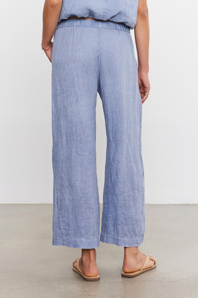 Woman standing in Velvet by Graham & Spencer's LOLA LINEN PANT ankle crop trousers and matching top, viewed from the back, with hands partially tucked in pockets.