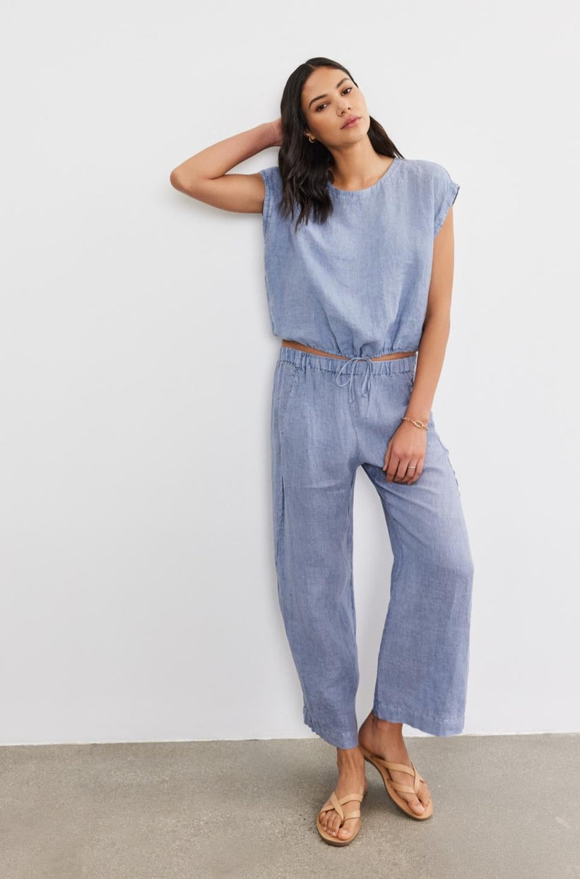Woman in blue Velvet by Graham & Spencer LOLA LINEN PANT trousers leaning against a white wall, looking at the camera.