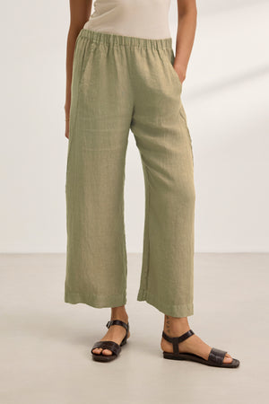 Wearing the Velvet by Graham & Spencer LOLA LINEN PANT, paired with a beige top and black sandals, a person stands on a light-colored floor against a plain background.
