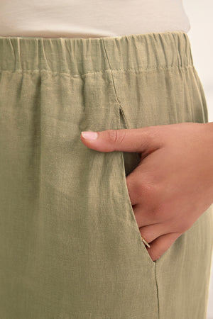 A close-up of a hand in the pocket of the LOLA LINEN PANT by Velvet by Graham & Spencer, featuring a lightweight linen fabric and elastic waistband in light olive green.