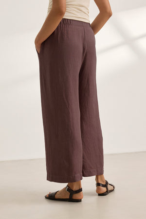Model wearing the LOLA LINEN PANT by Velvet by Graham & Spencer in brown with black sandals, standing hands-in-pockets against a plain, light background.