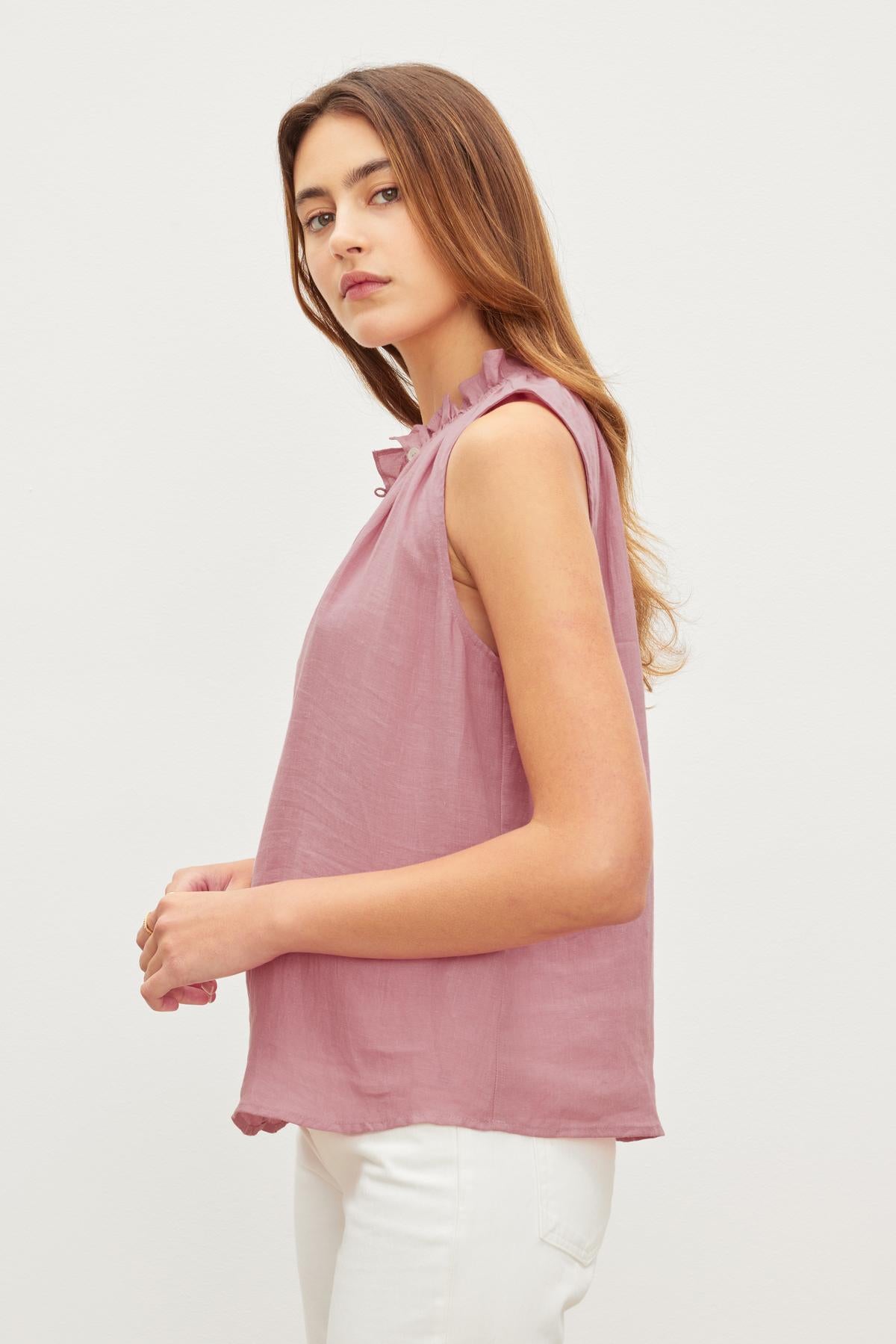 A woman with long brown hair in a pink Velvet by Graham & Spencer MASIE LINEN TANK TOP and white pants, standing side view with arms crossed.-36571197341889