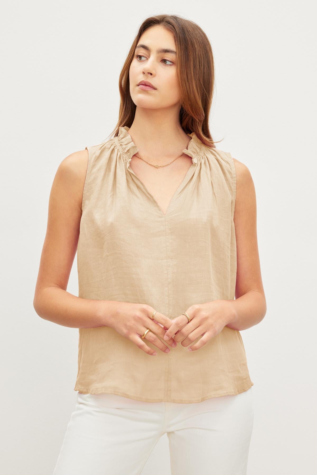 A woman wearing a sleeveless beige MASIE LINEN TANK TOP by Velvet by Graham & Spencer and white pants stands against a plain background.-37229082673345