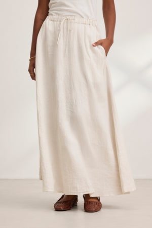 A person stands against a light background, wearing the long MEMPHIS LINEN SKIRT by Velvet by Graham & Spencer, featuring a cream color, woven linen fabric, elastic waist, and pockets. It is paired with brown shoes.
