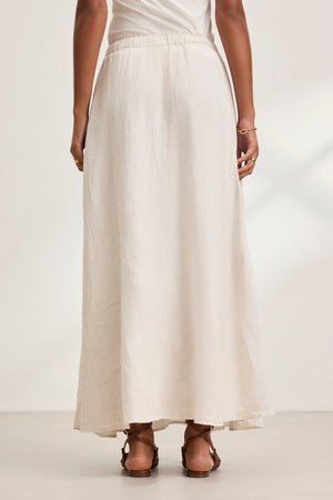 A person wearing the MEMPHIS LINEN SKIRT by Velvet by Graham & Spencer stands with their back to the camera, wearing a long, light beige woven linen maxi skirt with an elastic waist, paired with sandals and a white top.