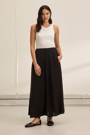 A woman indoors, dressed in a white sleeveless top and the flowing MEMPHIS LINEN SKIRT by Velvet by Graham & Spencer, strikes a pose with one hand in her pocket and pairs it with black shoes. The plain background enhances the elegance of her ensemble.